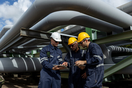 Engineer wearing safety gear using digital tablets at an industrial facility, discussing data and operations under large pipelines, symbolizing technology integration in modern engineering. - Powered by Adobe