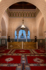 Interior view of the Mausoleum of Moulay Ismail in Meknes, Morocco, featuring ornate arches, intricate tilework, and colorful tapestries laid across the floor, illuminated by soft natural light.


