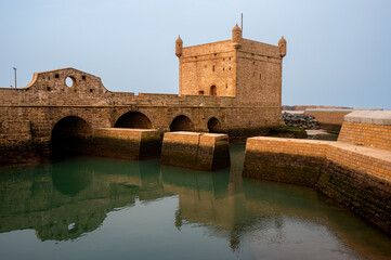 Wide golden hour shot of the stone bridge leading to the Castelo Real and the calm inner harbor of Essaouira, Morocco. The warm sunlight enhances the textures of the fort and water reflections.