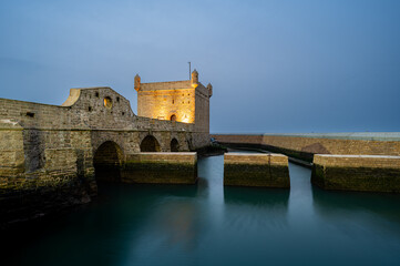 Wide evening long exposure shot of the stone bridge leading to the illuminated Castelo Real and the calm inner harbor of Essaouira, Morocco. The reflections of the lights shimmer across the harbor