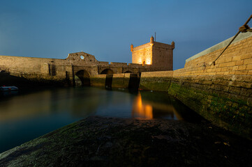 Long exposure night shot of the illuminated Castelo Real and the old harbor of Essaouira, Morocco. The glowing lights reflect softly on the calm water, creating a serene coastal night scene.