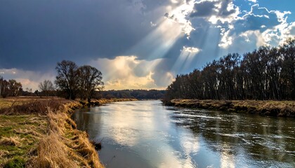 A serene river flows through a landscape with trees and golden grasses. Sunlight breaks through dramatic clouds