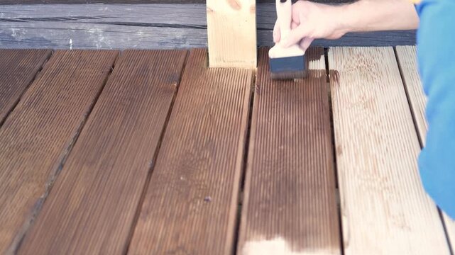 Brown oil for protection of wood by staining. Painting a larch for protecting and pests. Woodworker applying finish to terrace deck board. Repairman varnishing with brush wooden plank floor surface.