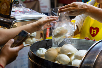 Close-up of a vendor handing a plastic bag with a steamed bun to a customer holding a smartphone at...