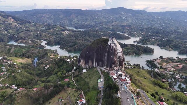 A cinematic, wide-angle circles the towering El Pe&ntilde;&oacute;n de Guatap&eacute; monolith in Colombia, capturing the massive rock formation, the expansive reservoir, and lush green islands during the daytime