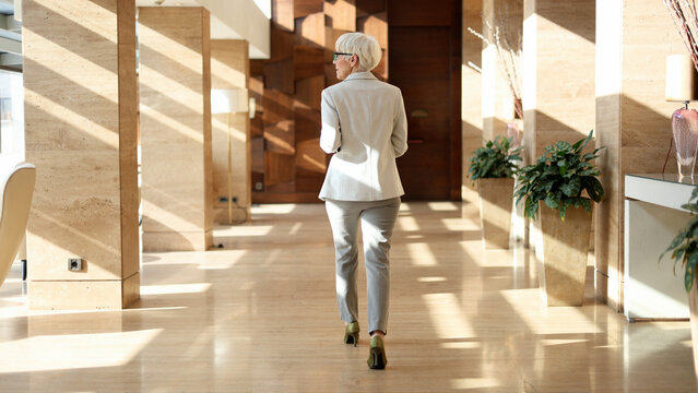 Mature businesswoman walking through modern office building corridor, facing away, career growth and corporate success