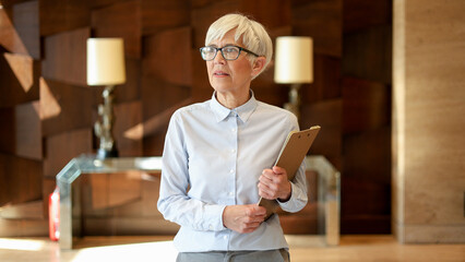 Senior businesswoman holding a clipboard in a modern office building, looking forward with focus and determination