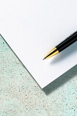 A black and gold pen is positioned on a white sheet of paper, which lies on a speckled, light blue and gray textured surface.
