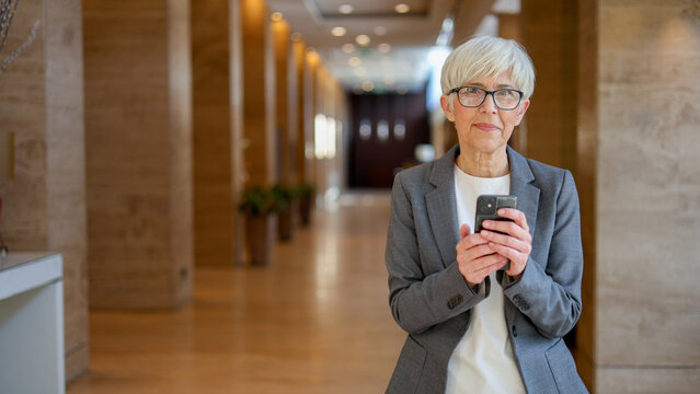 Senior businesswoman standing in a modern corporate hallway, holding smartphone and using mobile technology for communication