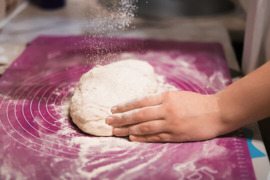A hand sprinkles flour onto a mound of fresh dough, which is being kneaded on a purple silicone baking mat.