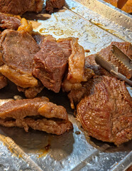 Overhead shot of several pieces of cooked, brown fried beef resting in a stainless steel tray with visible tongs on the side. Vertical view