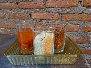 Three small glasses of common Asian condiments chili fish sauce, sugar, and dried chili flakes—arranged on a plastic tray against a weathered red brick wall.