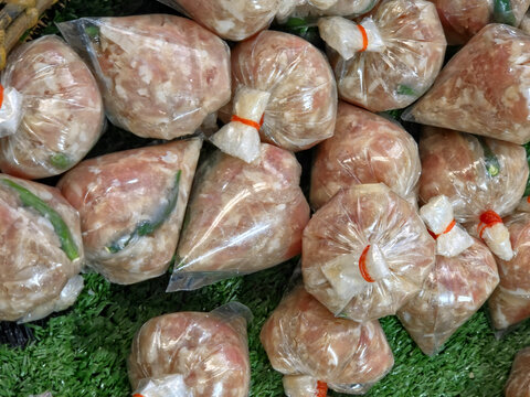 A close-up view of numerous individual portions of raw, light-pink ground meat (likely pork sausage or Isaan sour sausage) tightly wrapped and tied in clear plastic bags, resting on a green, textured 
