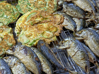 Close-up of a street food display with whole fried mackerel and vegetable-filled omelets on a metal grate.