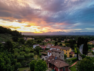 Aerial view of the Tuscan countryside during sunset, showing rustic village houses surrounded by olive groves, cypress trees, and rolling hills.