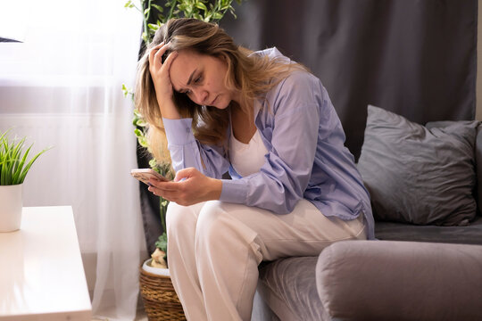 sad middle-aged woman sitting on couch at home reading message on her phone. A sad, tearful woman suffering from stress reads an emotional message.