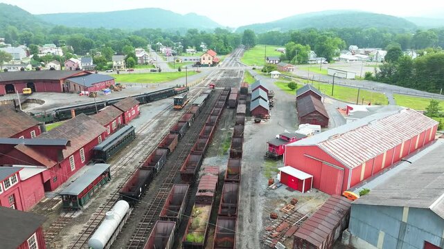 Aerial view of an active railway yard filled with train cars and surrounded by green hills and quaint buildings. The setting reflects a mix of industry and natural beauty.