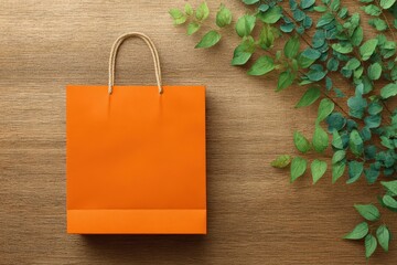 A vibrant orange shopping bag sits on a light brown wooden surface, complemented by a collection of fresh green leaves in a corner arrangement.