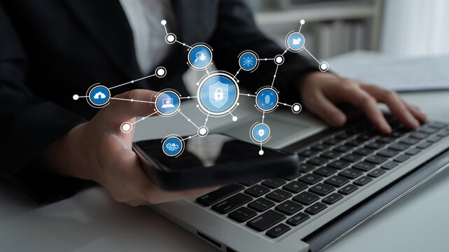 A business professional using a smartphone while working on a laptop, illustrating the intersection of technology and cybersecurity in a modern office environment. Tessel