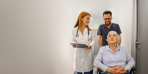 Male doctor walking beside coworker pushing senior patient on wheelchair discussing diagnosis in hospital corridor