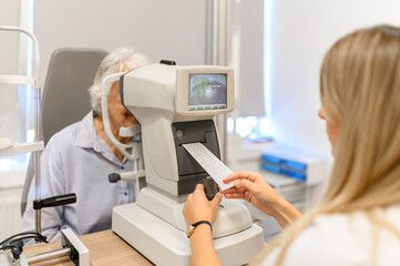 Female ophthalmologist observing paper printout from slit lamp while examining senior woman's eyes during vision test at clinic