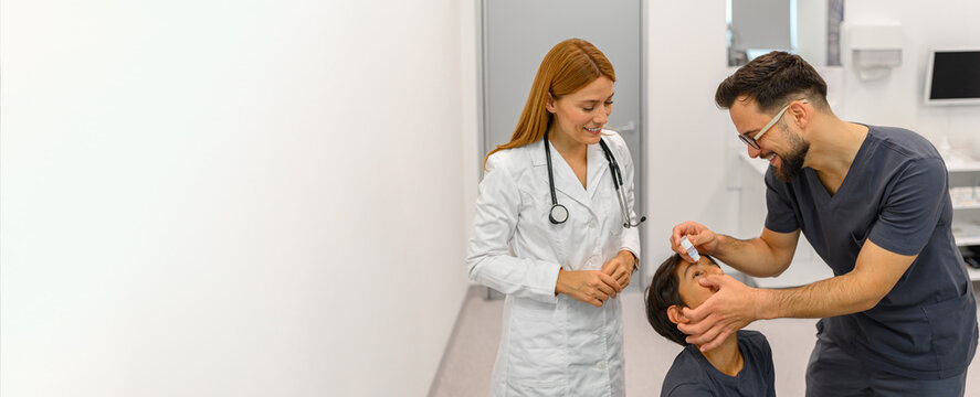 Woman looking at male doctor applying eye drops to boy's eye during hospital visit at ophthalmology clinic for vision checkup