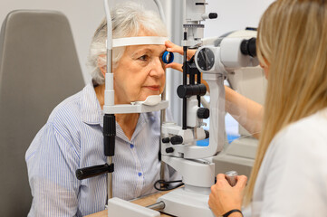 Female ophthalmologist examining senior woman's eyes with slit lamp while holding diagnostic lens...