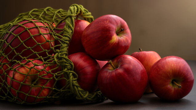 Ripe red apples spilling from a green reusable mesh bag. Still life with fresh fruit on a dark background. Healthy eating and sustainable shopping concept.