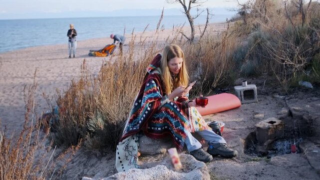 Young woman sitting near a fire pit on the beach, wrapped in a patterned blanket, holding a cup and smartphone. Friends prepare camping gear in the background. Cozy outdoor atmosphere.