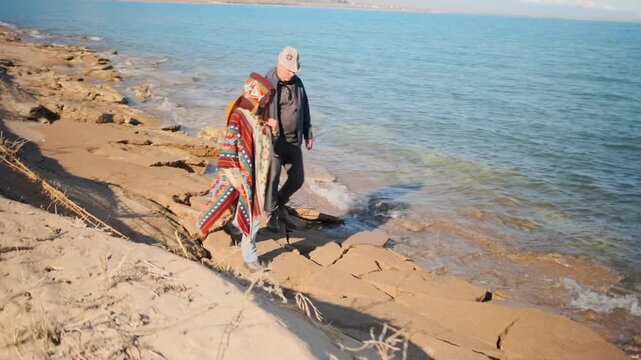 An older man and a young woman while walking on a rocky beach. Peaceful coastal atmosphere, gentle waves, quiet companionship, and warmth on a cool day.