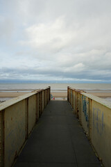 Rusty Pedestrian Bridge Leading To A Sandy Beach With The Sea And Offshore Wind Farm Under A Cloudy Sky In Towyn, North Wales, Uk.