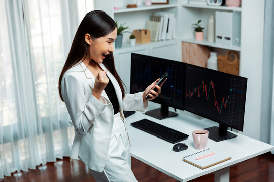 Successful young Asian businesswoman earning high profit raising fist up, looking smartphone for exchange stock market's financial technology with dynamic business graph at modern office. Stratagem.