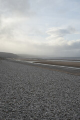 Scenic Pebble Beach And Sandy Tidal Flats With Misty Hills In The Background Under An Overcast Sky In Towyn, North Wales, Uk.