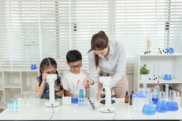 Caucasian female teacher teaching science using microscope with asian students in classroom full of beakers test tubes and blue liquid on table with bookshelves and white blinds in background