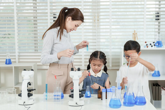 Asian boy and girl in elementary classroom performing hands-on science experiment using blue liquid and pipettes under supervision of smiling female caucasian teacher next to lab microscope