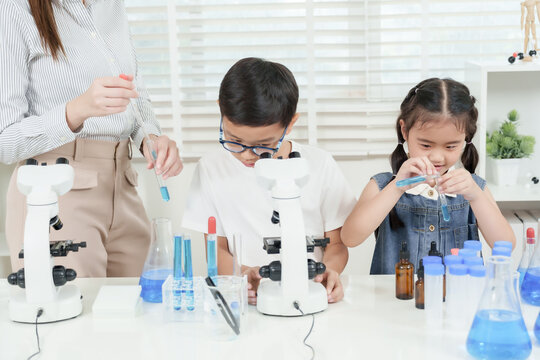 Asian elementary girl concentrating on mixing blue liquids in test tubes while working with classmates at science desk guided by female caucasian teacher inside bright classroom