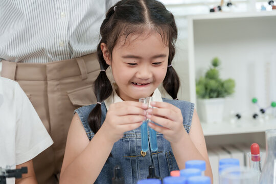 Asian elementary girl holding two test tubes smiling while comparing liquids during science class activity with microscope and lab equipment in background showing curiosity and enjoyment