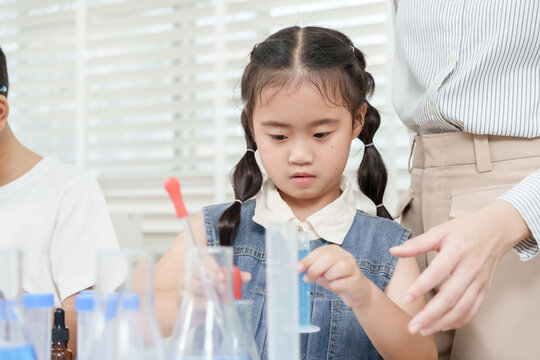 Multiracial elementary girl student learning science experiment with support of Asian female teacher while handling measuring cylinder at table with beakers, droppers and test tubes. - Powered by Adobe