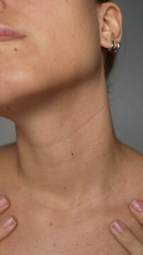 Vertical video of a young woman touching her neck with her hands on a light grey background. Lines on the female neck. Wrinkles, age-related changes, rings of Venus, creases