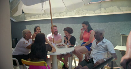 Diverse group of friends and family seated around table sharing laughs and enjoying food at outdoor backyard barbecue, celebrating joy and togetherness in summer