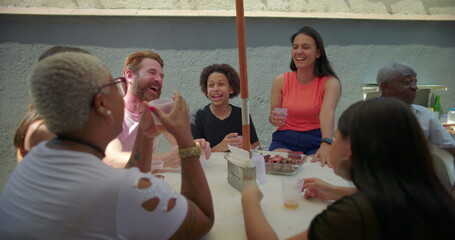Diverse group of friends and family seated around table sharing laughs and enjoying food at outdoor backyard barbecue, celebrating joy and togetherness in summer