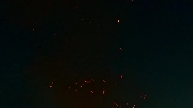 Bright glowing orange sparks scatter in darkness and drift upward against a black background creating dynamic fiery motion reminiscent of burning embers at night