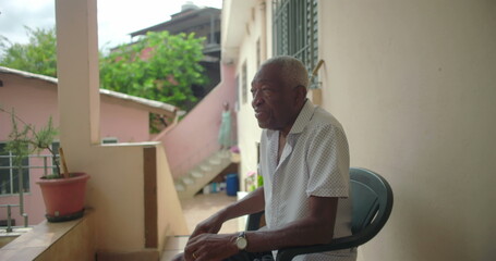 One Elderly Black man seated outdoors on porch, gazing thoughtfully, surrounded by warm tropical architecture, pink walls, serene and contemplative expression