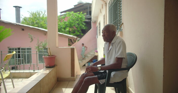 One Elderly Black man seated outdoors on porch, gazing thoughtfully, surrounded by warm tropical architecture, pink walls, serene and contemplative expression
