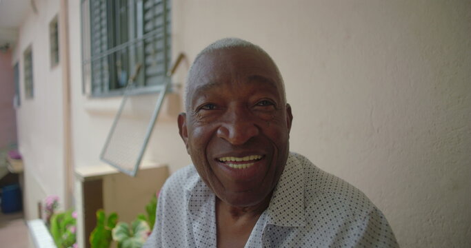 One Elderly Black man smiling warmly while seated outdoors on a porch, exuding joy and positivity, with tropical residential surroundings in the background - Powered by Adobe