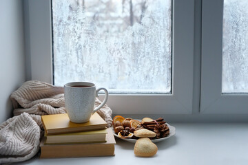 still life with cup of tea or coffee, books, cookies, nuts in plate, sweater on windowsill. frozen window background. home hygge comfort in cold weather. winter season. Christmas, New Year holidays