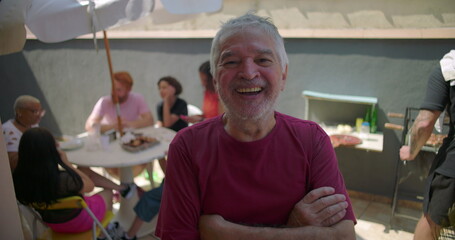 Elderly man smiling warmly at a backyard barbecue, surrounded by a lively gathering of family and friends enjoying food and connection in an outdoor celebration