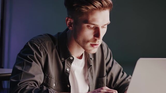A young man types intently on a laptop, focusing on the task at hand in a dimly lit room. The man&rsquo;s attention to the laptop reflects his dedication to completing important work.