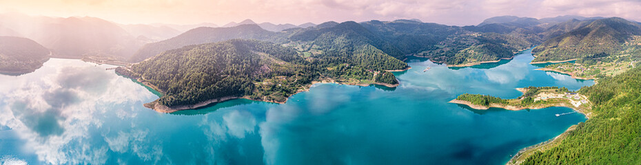 Aerial view of a stunning turquoise lake curving through lush green mountains and forests, reflecting clouds on the water surface