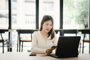 Fototapeta premium Young woman using a credit card to make online payments on her laptop in a modern cafe, representing digital lifestyle and e-commerce convenience.
